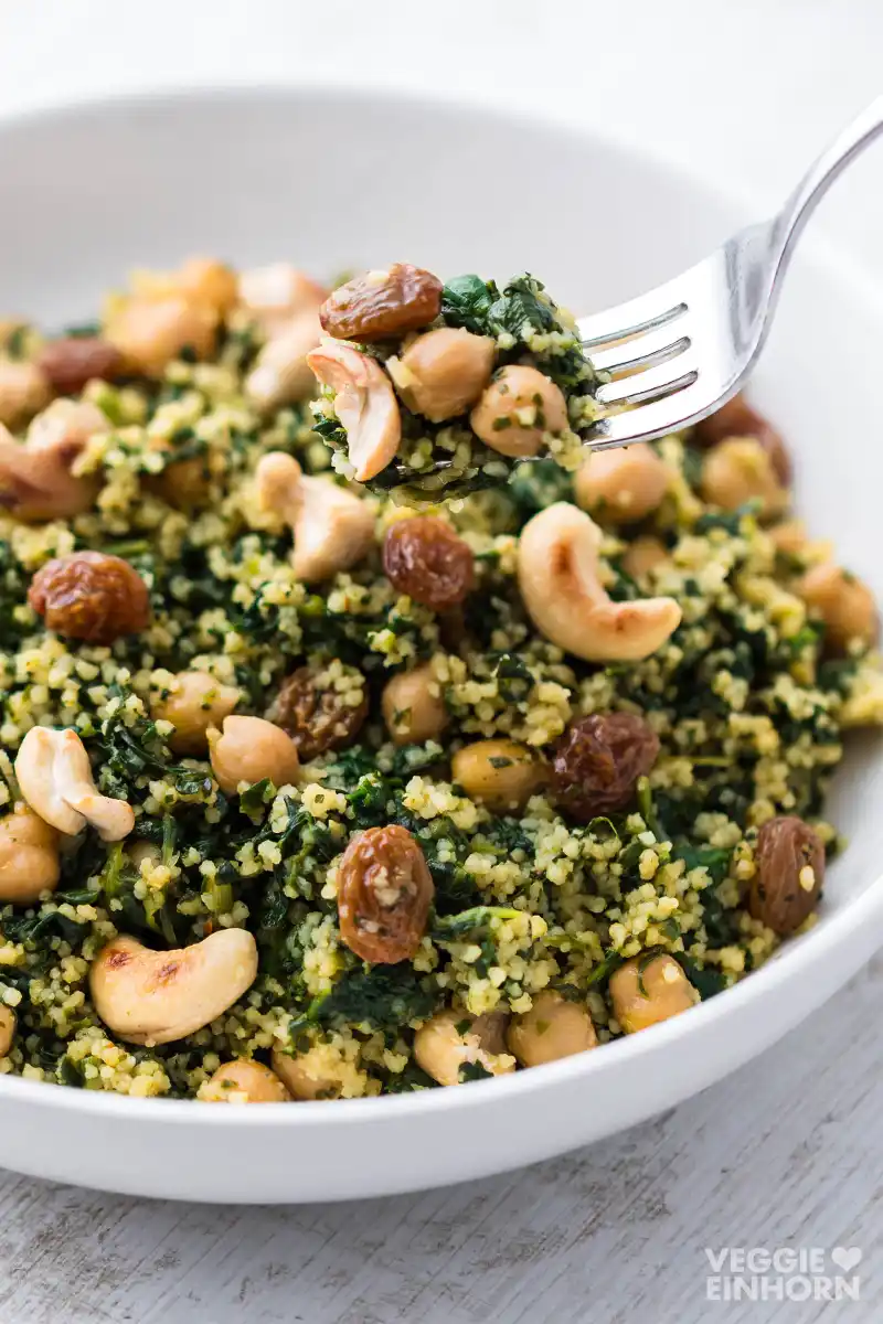 Weißer Teller mit einer Portion Couscous-Pfanne mit Spinat, Kichererbsen, Cashewkernen und Rosinen, eine Gabel hebt etwas von dem Gericht an.