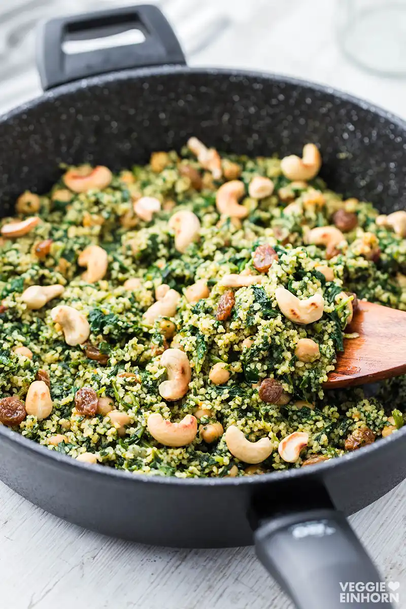 Nahaufnahme einer schwarzen Pfanne mit veganer Couscous-Pfanne, darin Spinat, Kichererbsen, geröstete Cashewkerne, Rosinen und ein Holzlöffel.