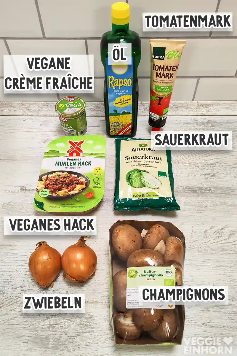 Verschiedene Zutaten wie veganes Hack, Sauerkraut, Champignons, Zwiebeln, Öl, Tomatenmark und vegane Crème fraîche liegen auf einer hellen Arbeitsfläche.