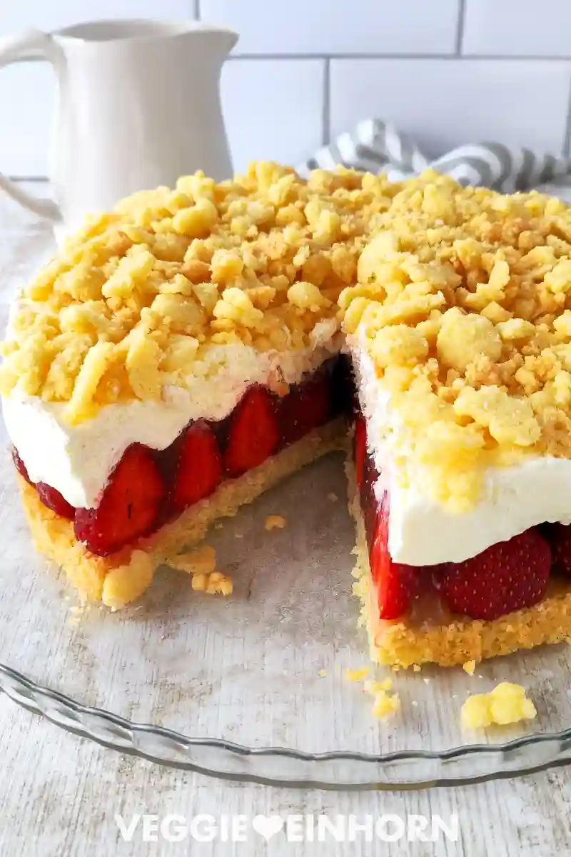 Vegane Erdbeertorte mit Sahneschicht, frischen Erdbeeren und goldgelben Streuseln auf einer Glas-Tortenplatte serviert.