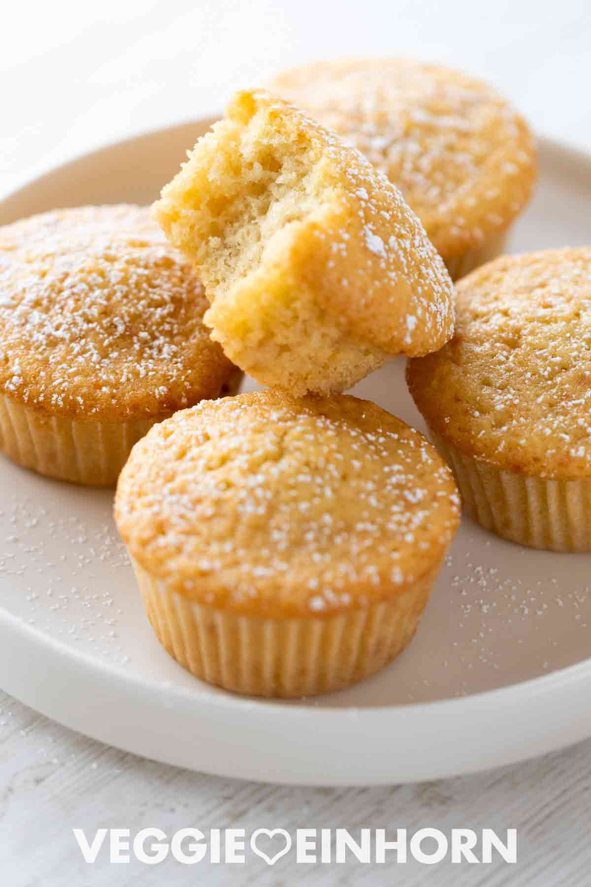 Mehrere goldbraune vegane Muffins in Papierförmchen, mit Puderzucker bestäubt, auf einem weißen Teller. Ein Muffin ist angebissen, sodass die fluffige Textur sichtbar ist.
