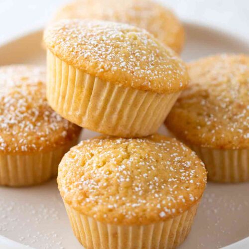Nahaufnahme von veganen Muffins in Papierförmchen, mit Puderzucker bestäubt, auf einem weißen Teller.