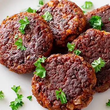Nahaufnahme von mehreren veganen Frikadellen aus Kidneybohnen, Linsen und Sojagranulat auf einem weißen Teller, garniert mit frischer Petersilie.