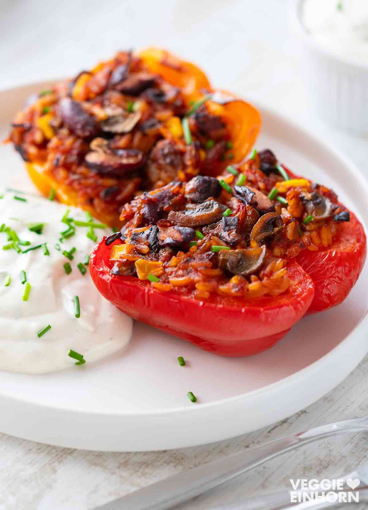 Gefüllte Paprika mit einer veganen Mischung aus Reis, Kidneybohnen, Mais und Champignons, serviert auf einem weißen Teller mit veganem Kräuterquark und frischem Schnittlauch, im Hintergrund ein kleines Schälchen.