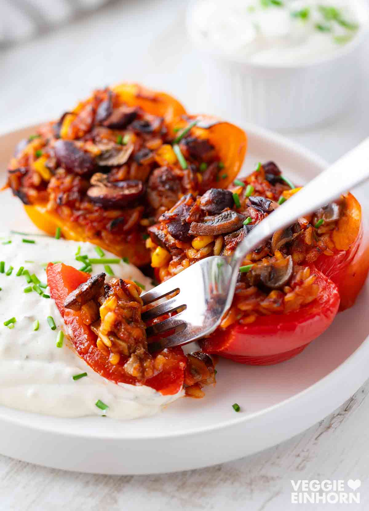 Nahaufnahme von gefüllten Paprika mit Reis, Kidneybohnen, Mais und Champignons, eine Gabel nimmt ein Stück auf. Daneben ein veganer Kräuterquark.