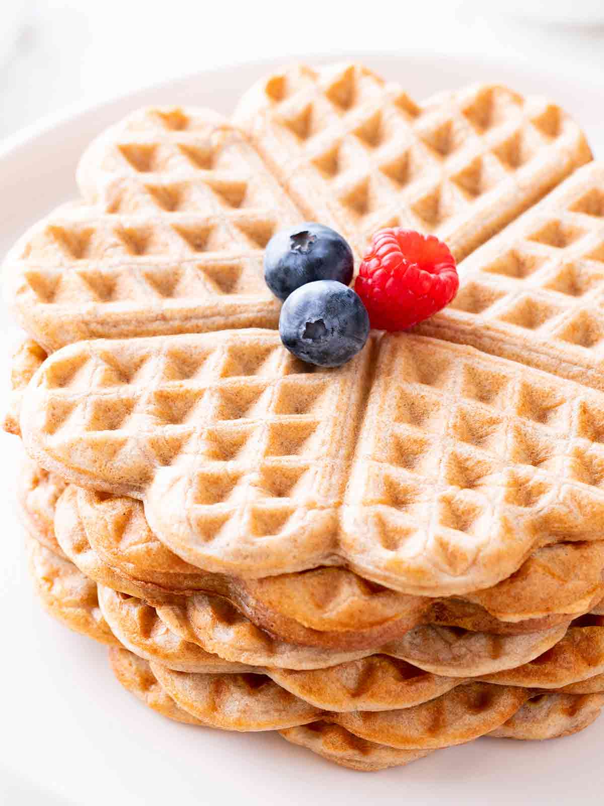Nahaufnahme von herzförmigen veganen Vollkornwaffeln ohne Zucker, mit frischen Beeren dekoriert, auf einem weißen Teller serviert.