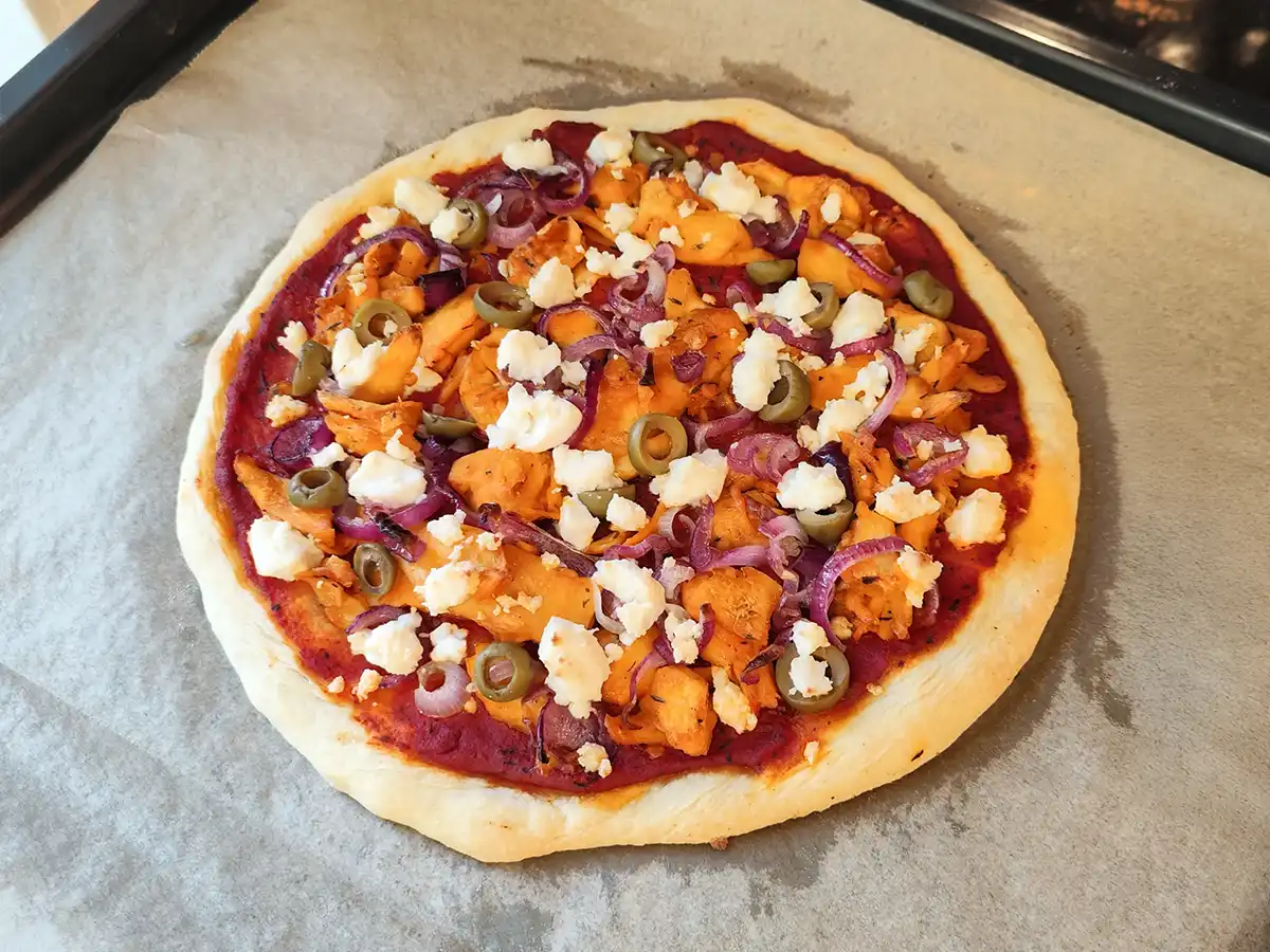 Vegane Gyrospizza auf einem Backblech mit Tomatensauce, veganen Gyrosstücken, roten Zwiebeln, Oliven und zerbröckeltem veganem Feta.