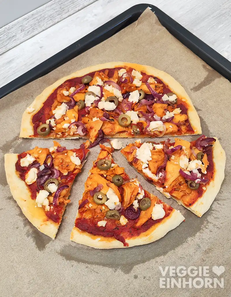 In Stücke geschnittene vegane Gyrospizza mit Pizzasauce, veganem Gyros, grünen Oliven, roten Zwiebeln und veganem Feta auf einem Backblech mit Backpapier.