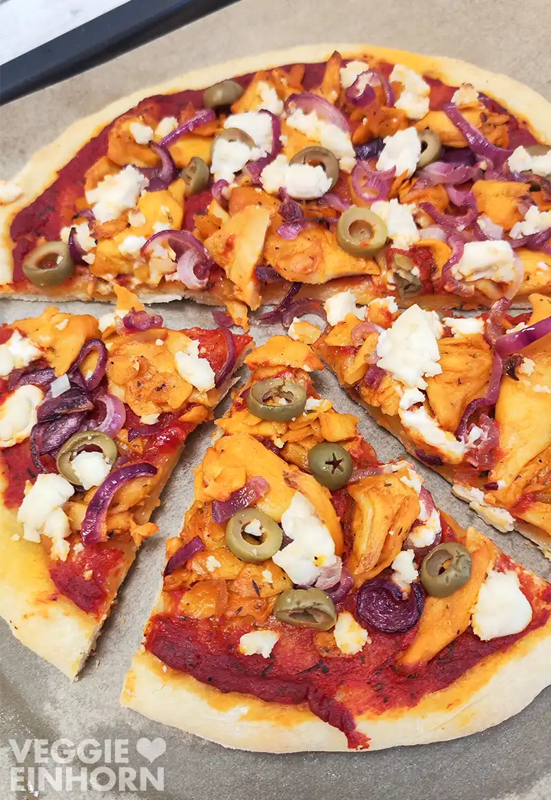 Nahaufnahme einer in Stücke geschnittenen veganen Pizza mit veganem Gyros, veganem Feta, grünen Olivenscheiben und roten Zwiebelringen.