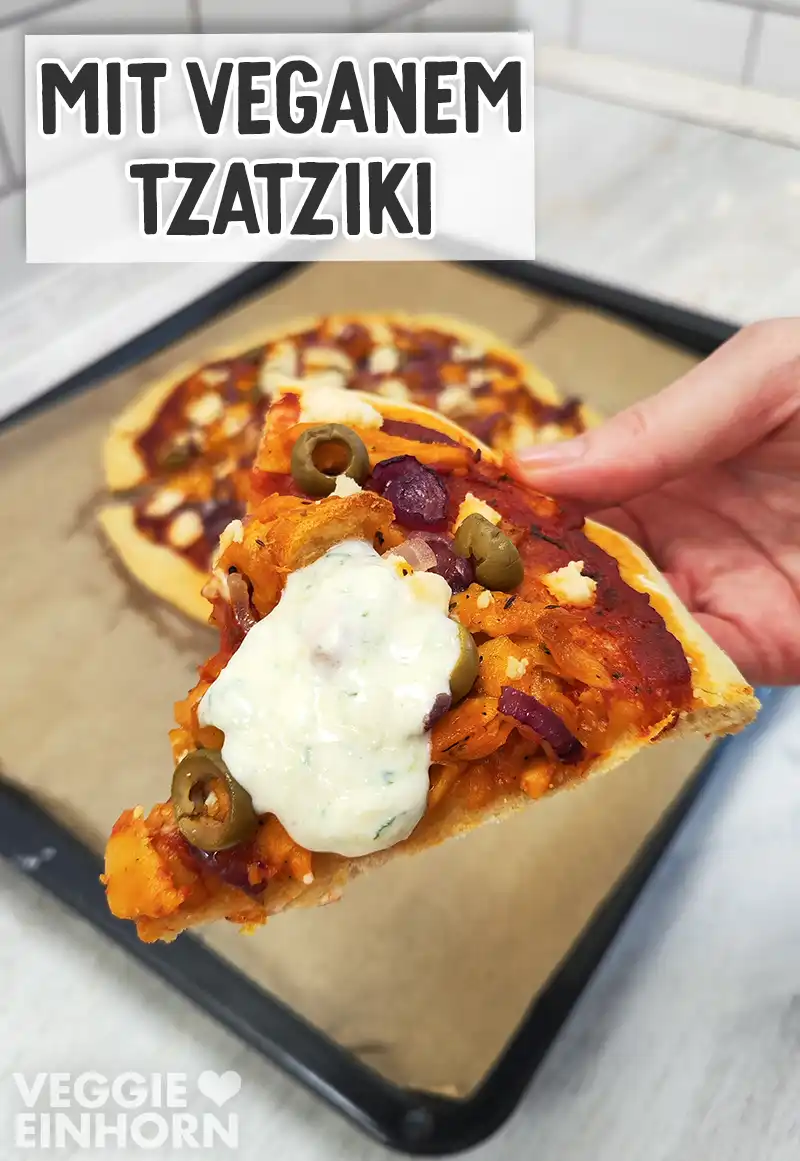 Eine Hand hält ein Stück vegane Gyrospizza mit Oliven, roten Zwiebeln und veganem Feta, auf dem Pizzastück ist ein Klecks veganer Tzatziki.
