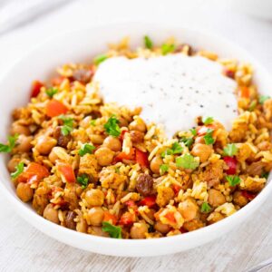 Weißer Teller mit veganer Hackfleisch-Reis-Pfanne aus Kichererbsen, Paprika, veganem Hack, Rosinen, Reis und Joghurt auf einem weißen Tisch.