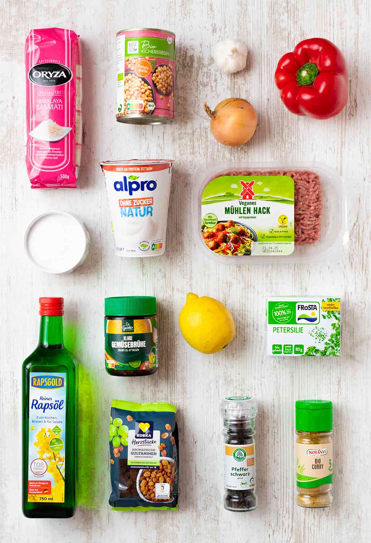 Verpackungen und Zutaten für eine vegane Hackfleisch-Reis-Pfanne auf einem weißen Holzuntergrund: Reis, Kichererbsen, Paprika, veganes Hack, Joghurt, Zitrone, Rosinen, Öl, Gewürze und Kräuter.