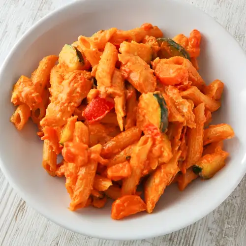 Cremige vegane Hähnchenpfanne mit Penne, Zucchini, Möhren und Tomaten auf einem weißen Teller.