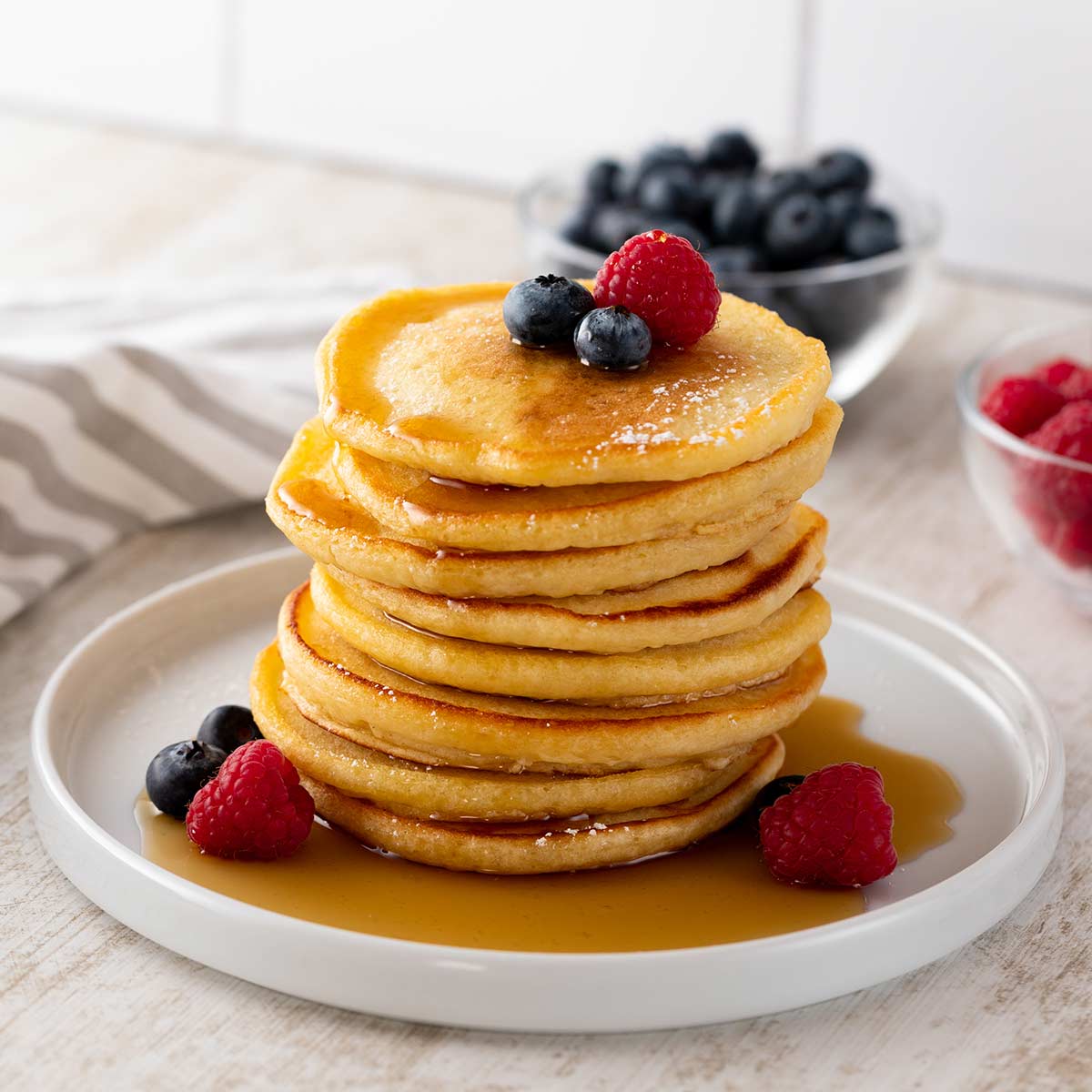 Ein Stapel fluffiger veganer Pancakes, serviert mit Ahornsirup, Puderzucker und frischen Beeren, auf einem weißen Teller.