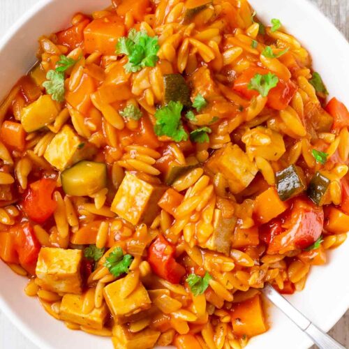 Nahaufnahme einer veganen Kritharaki-Pfanne mit Räuchertofu, Paprika, Zucchini und Möhren in einer aromatischen Tomatensoße, serviert in einem weißen Teller und garniert mit frischer Petersilie.