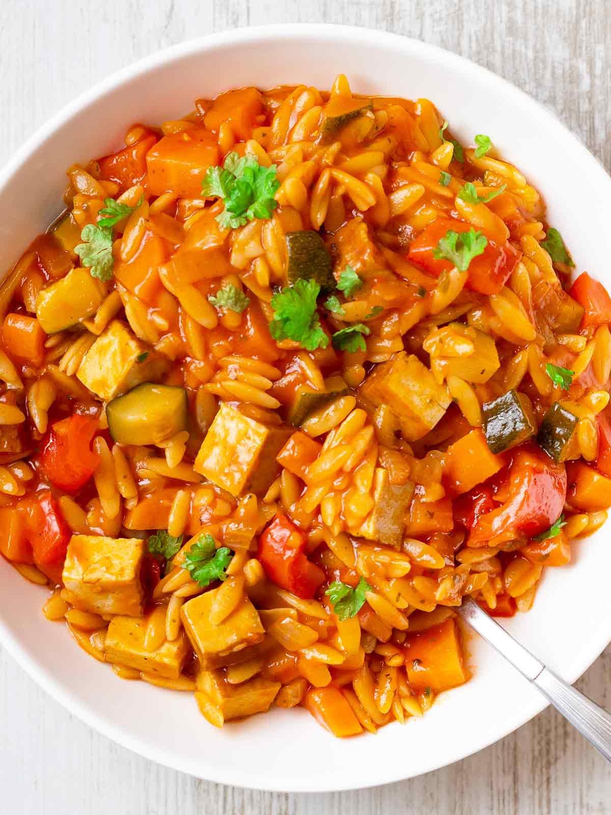 Nahaufnahme einer veganen Kritharaki-Pfanne mit Räuchertofu, Paprika, Zucchini und Möhren in einer aromatischen Tomatensoße, serviert in einem weißen Teller und garniert mit frischer Petersilie.