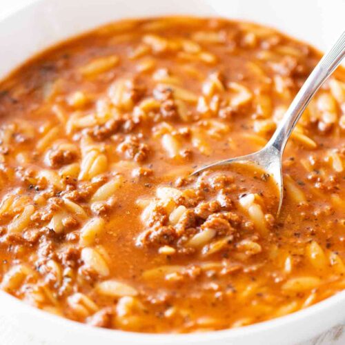 Eine weiße Schüssel mit rötlich-brauner Kritharaki-Suppe, gefüllt mit kleinen Orzo-Nudeln und krümeligem veganem Hack. Ein Löffel ist teilweise in die Suppe eingetaucht.