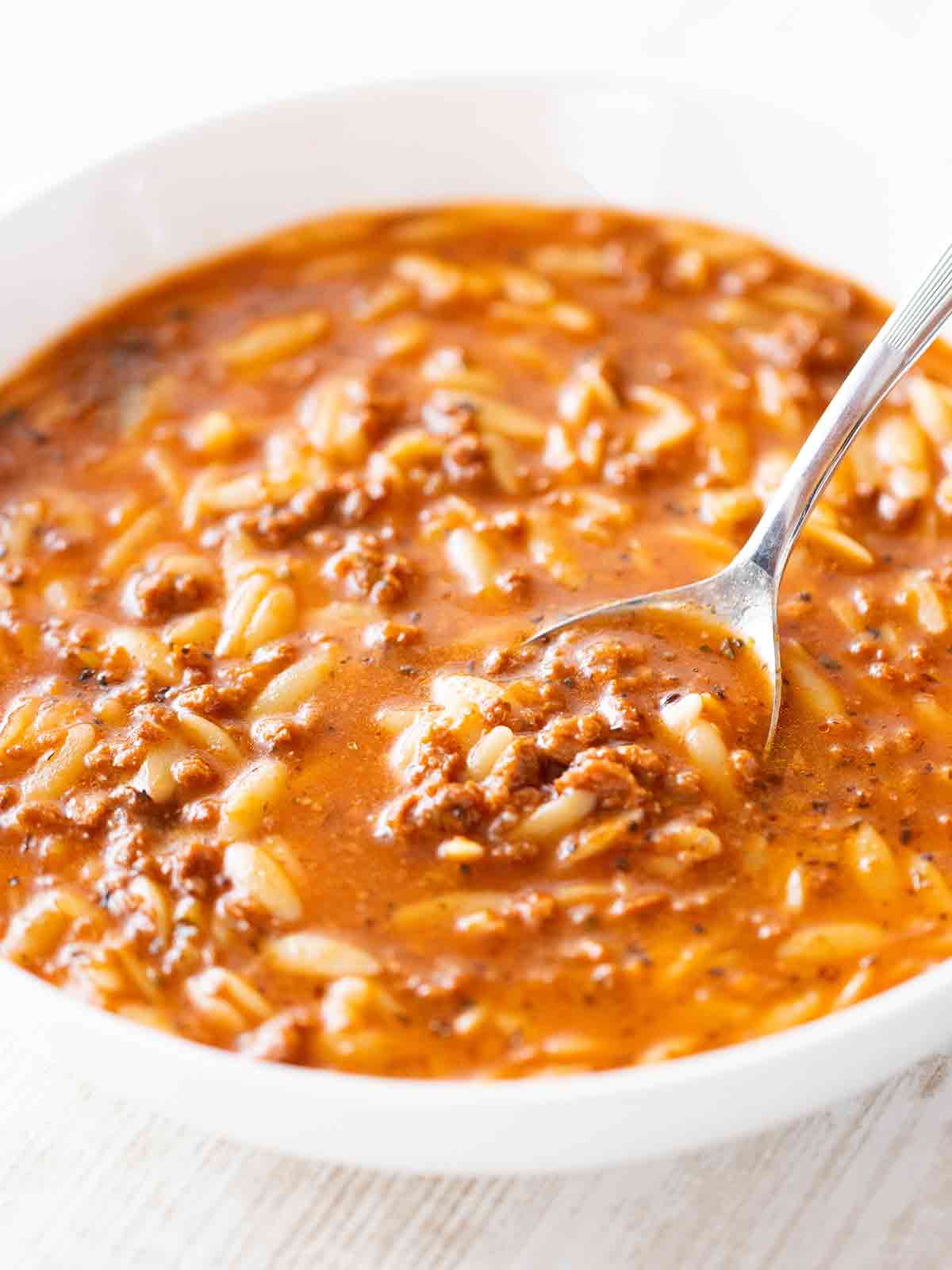 Eine weiße Schüssel mit rötlich-brauner Kritharaki-Suppe, gefüllt mit kleinen Orzo-Nudeln und krümeligem veganem Hack. Ein Löffel ist teilweise in die Suppe eingetaucht.