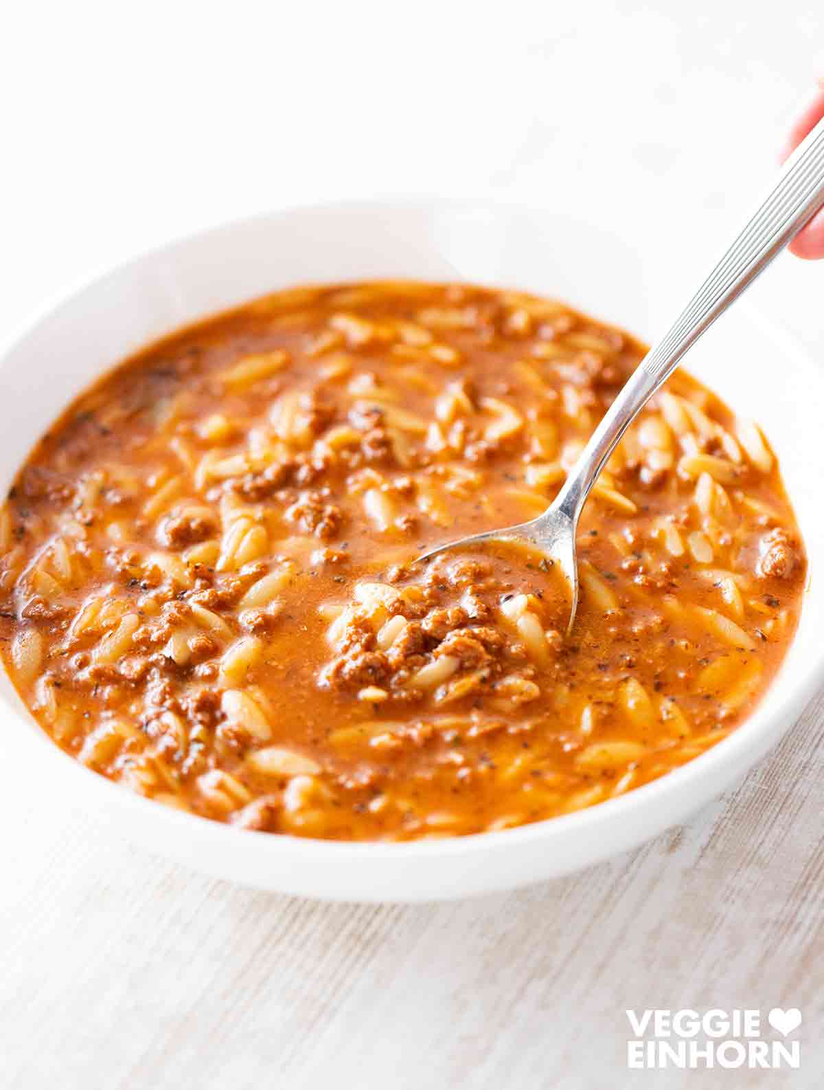Eine Schüssel mit Kritharaki-Suppe, in der kleine Orzo-Nudeln und krümeliges veganes Hack zu sehen sind. Eine Hand hält einen Löffel, der in die Suppe eintaucht. 