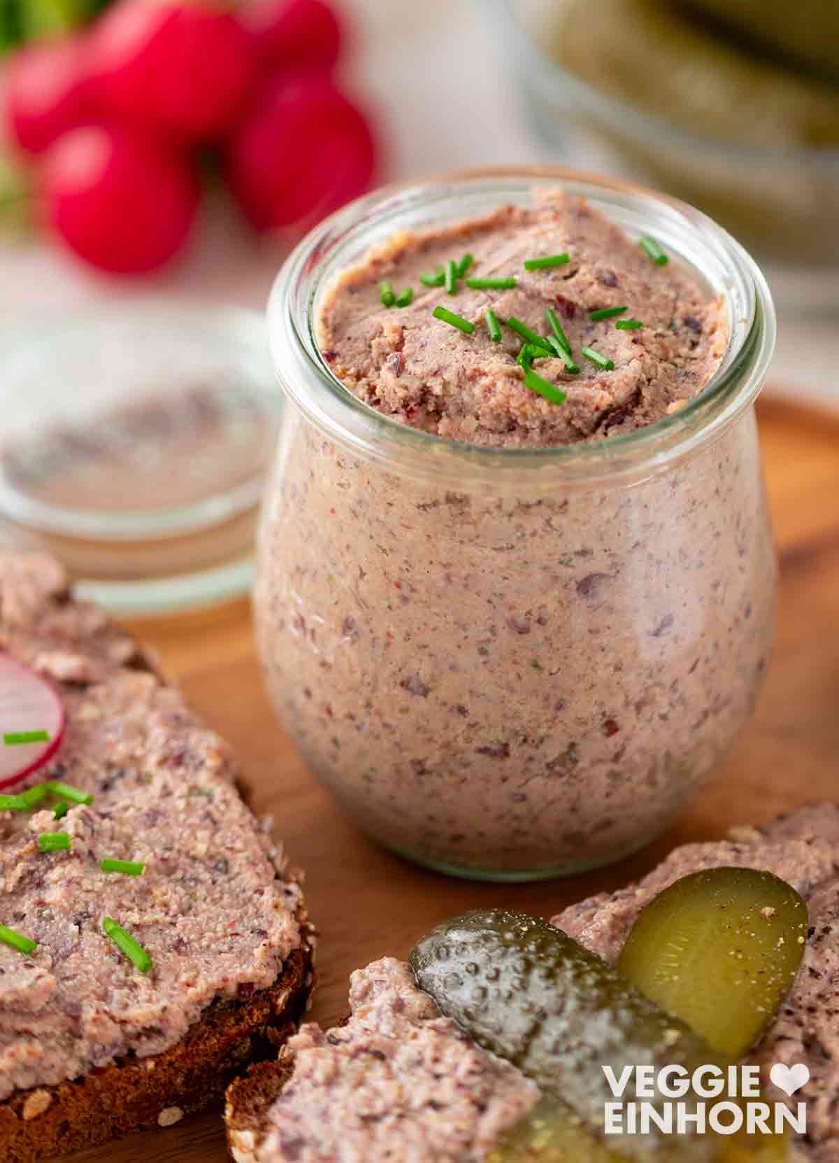 Ein Glas mit veganer Leberwurst, garniert mit frischem Schnittlauch. Daneben eine Scheibe Vollkornbrot mit der Streichwurst, belegt mit Radieschenscheiben und Schnittlauch. Im Vordergrund eine weitere Scheibe Brot mit veganer Leberwurst und zwei Essiggurkenscheiben. Im Hintergrund frische Radieschen.