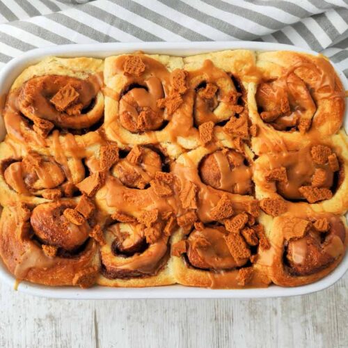 Vegane Lotus Zimtschnecken in einer weißen Form, goldbraun gebacken und mit karamellfarbener Biscoff Glasur überzogen. Obenauf liegen knusprige Lotuskeks Stückchen.