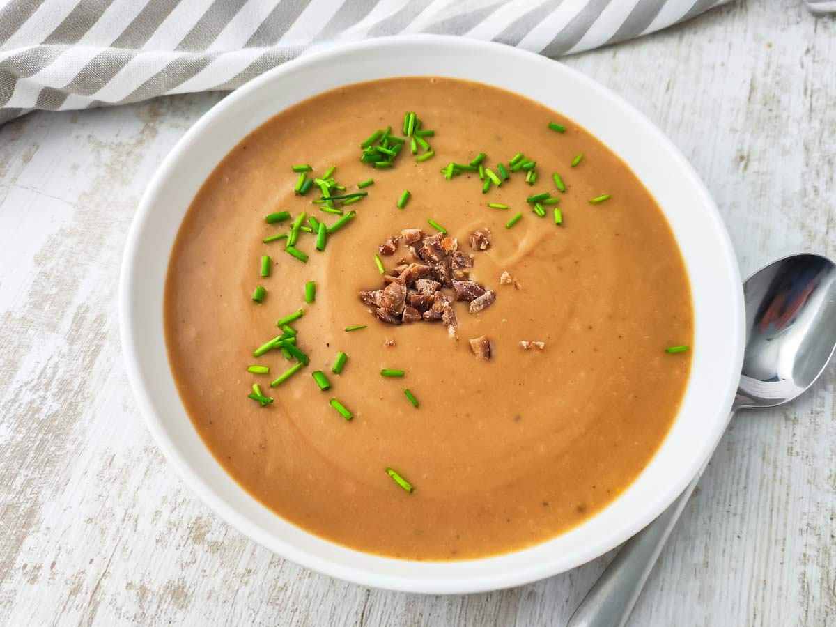 Vegane Maronen-Kartoffelsuppe in einem weißen Teller mit grünen Schnittlauchringen und gehackten Maronen, daneben ein Löffel auf hellem Holzuntergrund.