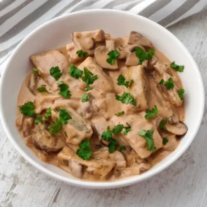 Teller mit veganen Maultaschen in Champignon Rahmsoße mit Petersilie auf hellem Holztisch.