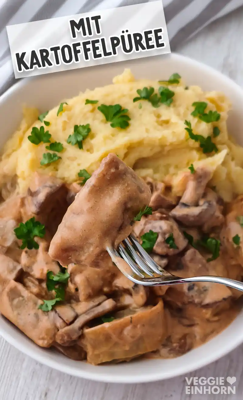 Gabel mit veganer Maultasche vor einem Teller mit weiteren Maultaschen, Champignon Rahmsoße und Kartoffelpüree.