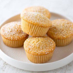Fluffige vegane Muffins in Papierförmchen, mit Puderzucker bestäubt, auf einem weißen Teller.