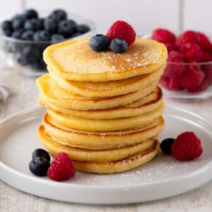 Ein Stapel goldbrauner veganer Pancakes, garniert mit frischen Himbeeren und Blaubeeren, serviert auf einem weißen Teller. Im Hintergrund befinden sich Schalen mit zusätzlichen Beeren.