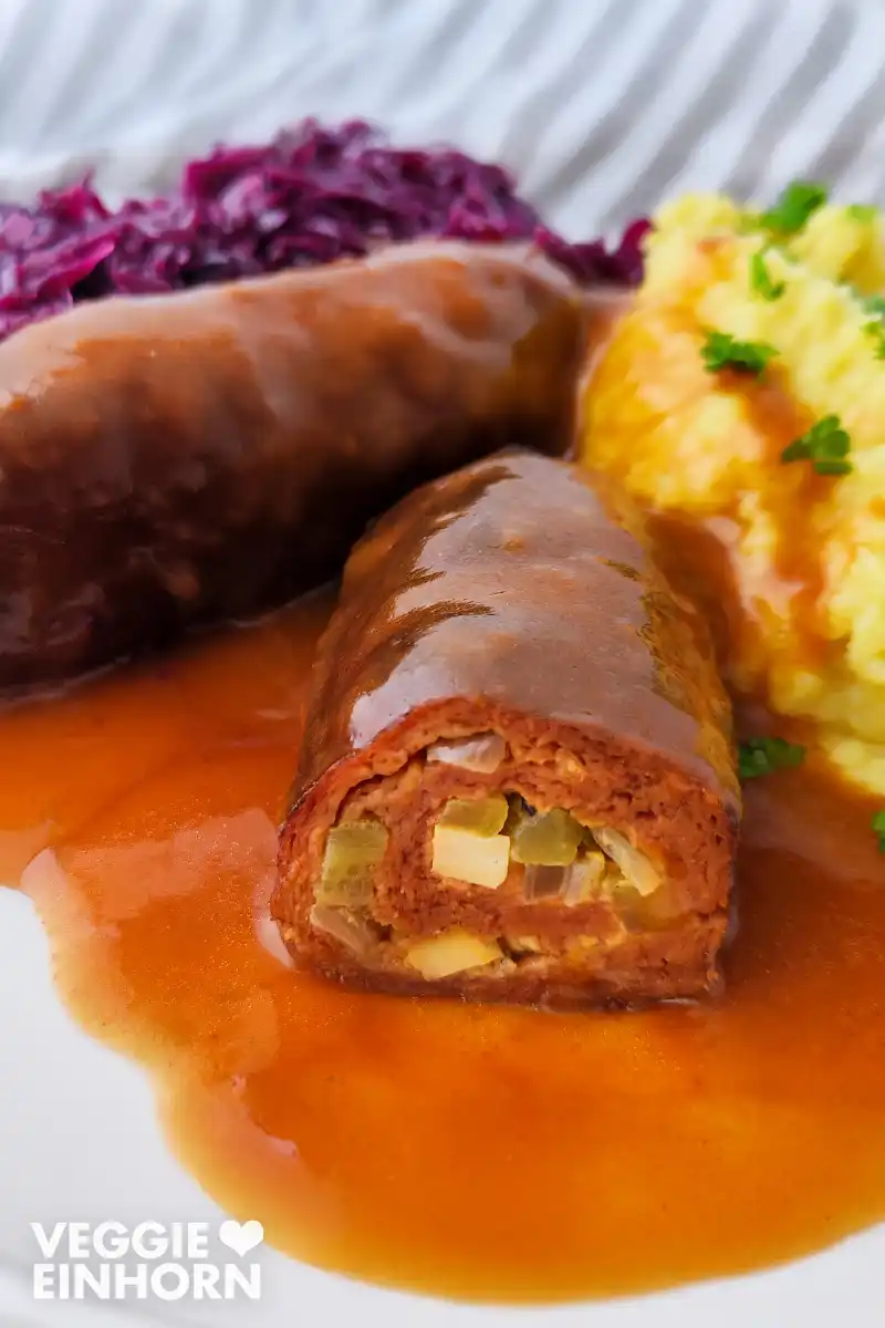 Nahaufnahme einer aufgeschnittenen veganen Seitanroulade in brauner Soße mit sichtbarer Füllung, serviert mit Kartoffelpüree und Rotkohl auf einem weißen Teller. Im Hintergrund liegt eine zweite Roulade.