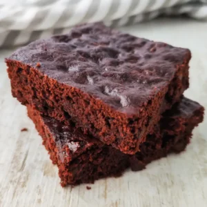 Zwei vegane Schoko Brownies liegen übereinander auf einem hellen Holztisch mit einem gestreiften Tuch im Hintergrund.