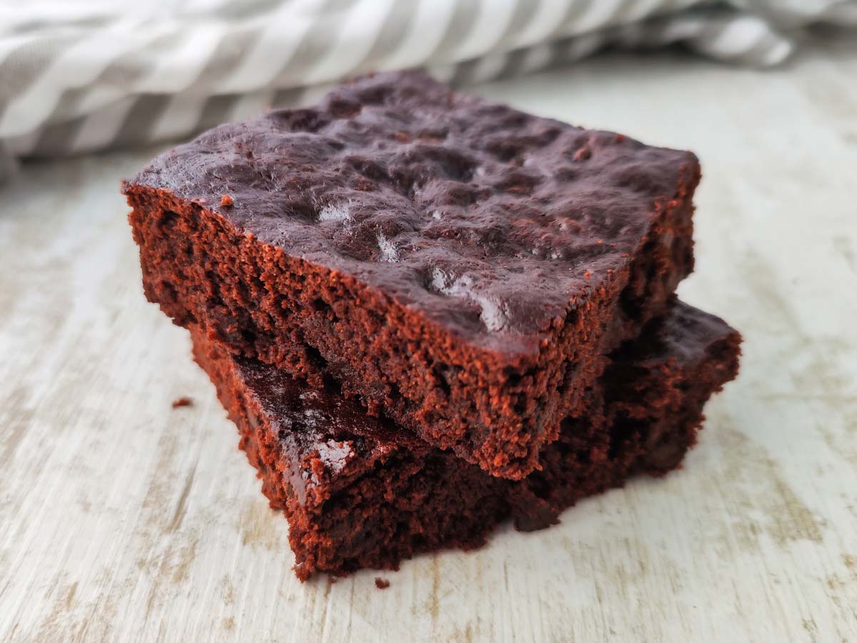 Zwei übereinander gestapelte vegane Schokobrownies auf heller Holzoberfläche mit gestreiftem Tuch im Hintergrund.