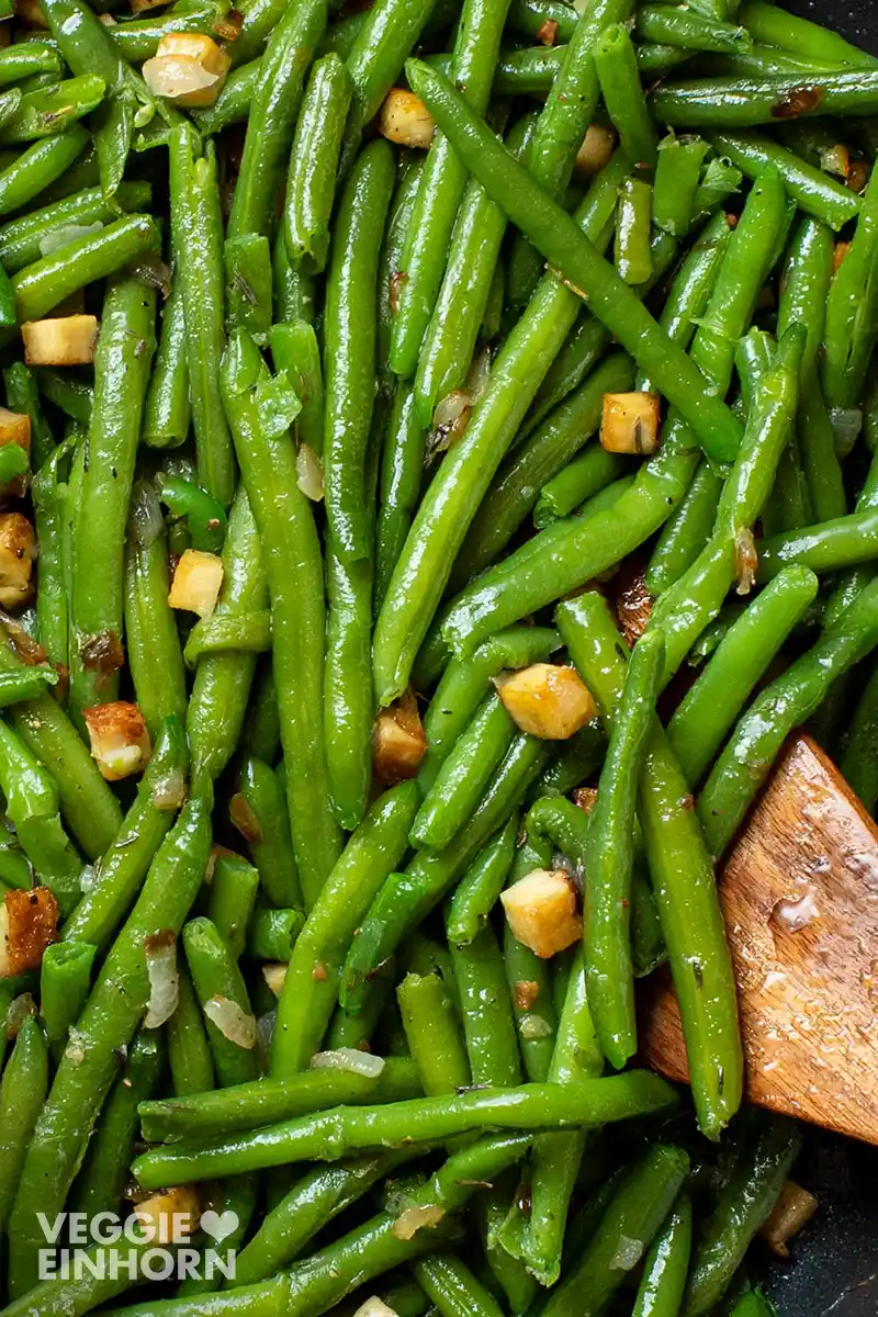 Nahaufnahme von grünen Bohnen mit Räuchertofu-Würfeln und einem Pfannenwender aus Holz.
