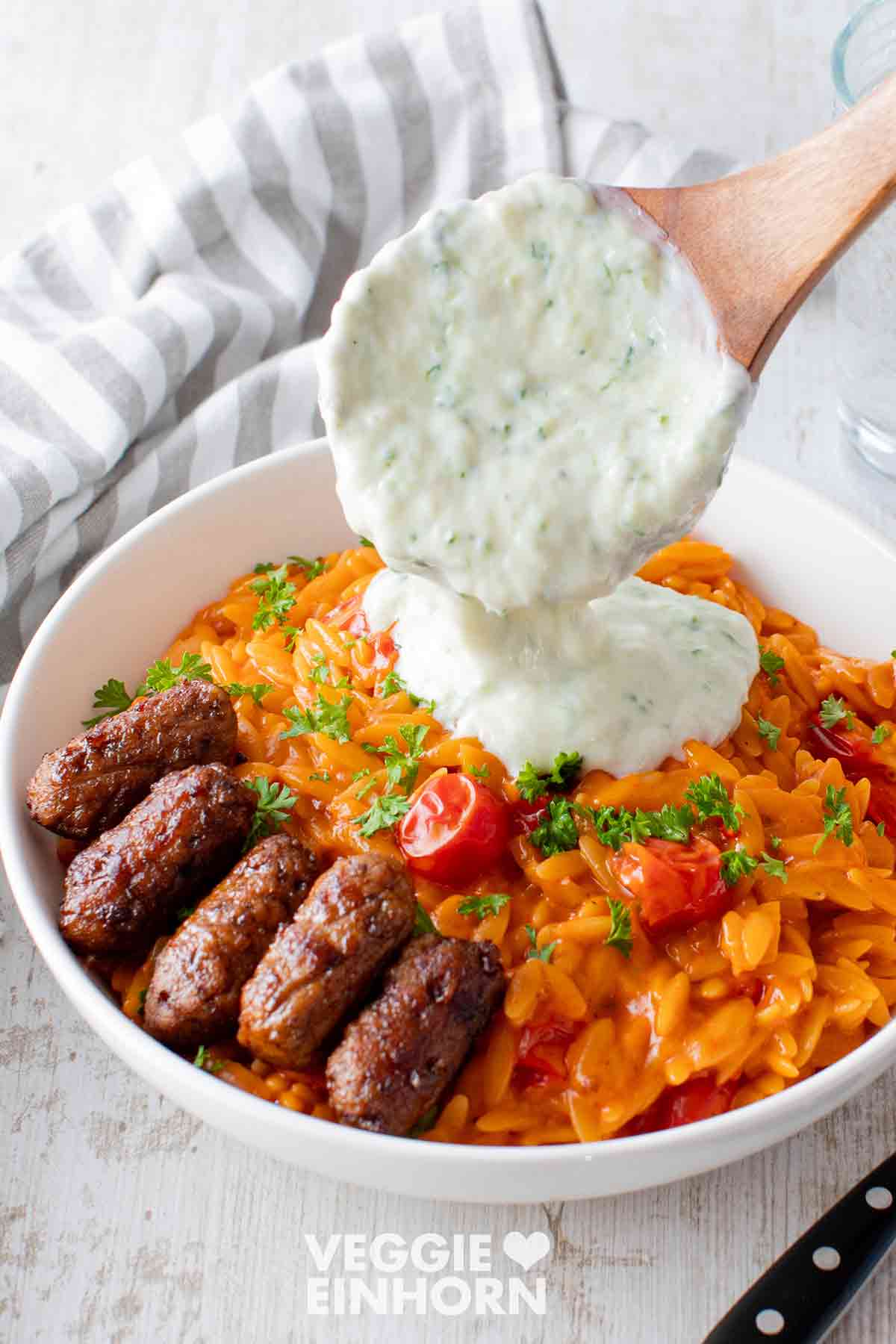 Veganer Tzatziki wird auf den Teller mit Tomaten Kritharaki und veganen Cevapcici gegeben
