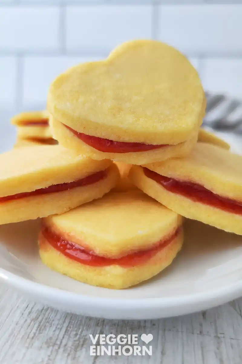 Vegane Vanilleplätzchen mit Marmeladenfüllung Gestapelte herzförmige vegane Vanilleplätzchen mit einer dünnen Schicht roter Marmelade dazwischen auf einem weißen Teller.