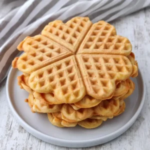 Mehrere herzförmige vegane Waffeln liegen gestapelt auf einem grauen Teller auf einem hellen Holztisch, im Hintergrund liegt ein gestreiftes Küchentuch.