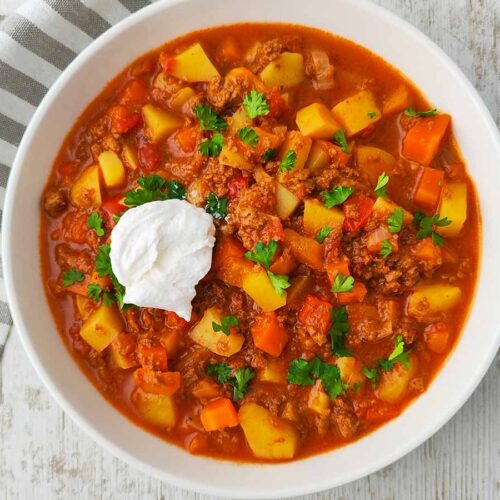 Teller mit veganem Bauerntopf mit veganem Hackfleisch, Kartoffeln, Möhren und Paprika auf hellem Holztisch.
