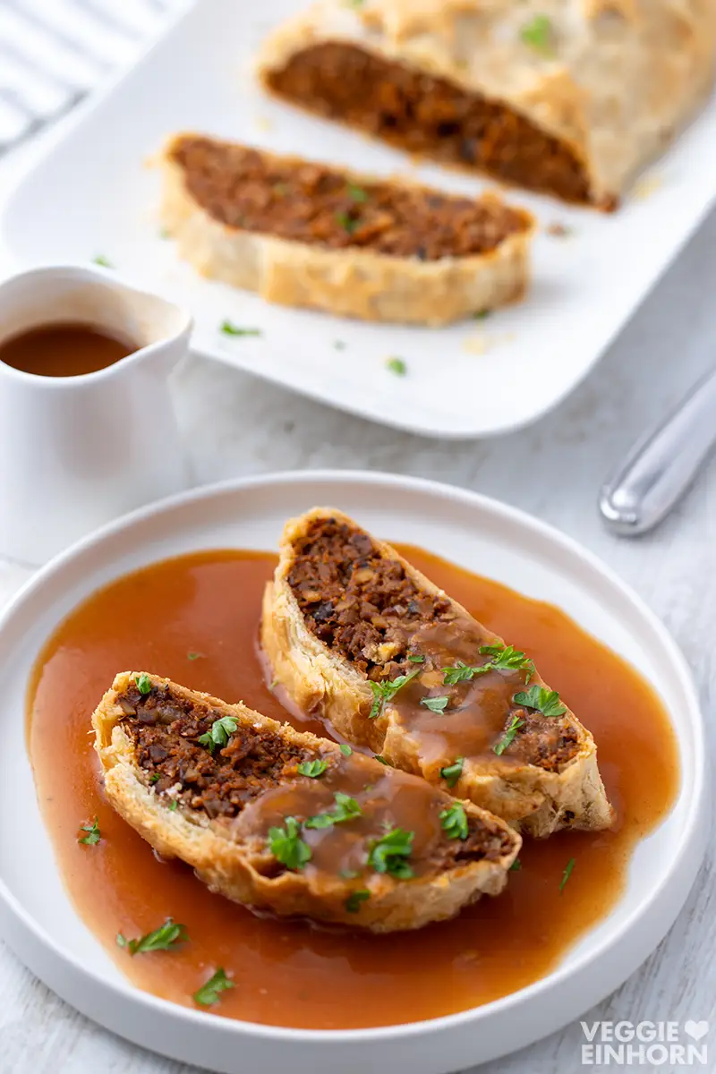 Aufgeschnittener veganer Braten im Blätterteig mit Sauce auf einem Teller, im Hintergrund der restliche Braten auf einer Servierplatte.