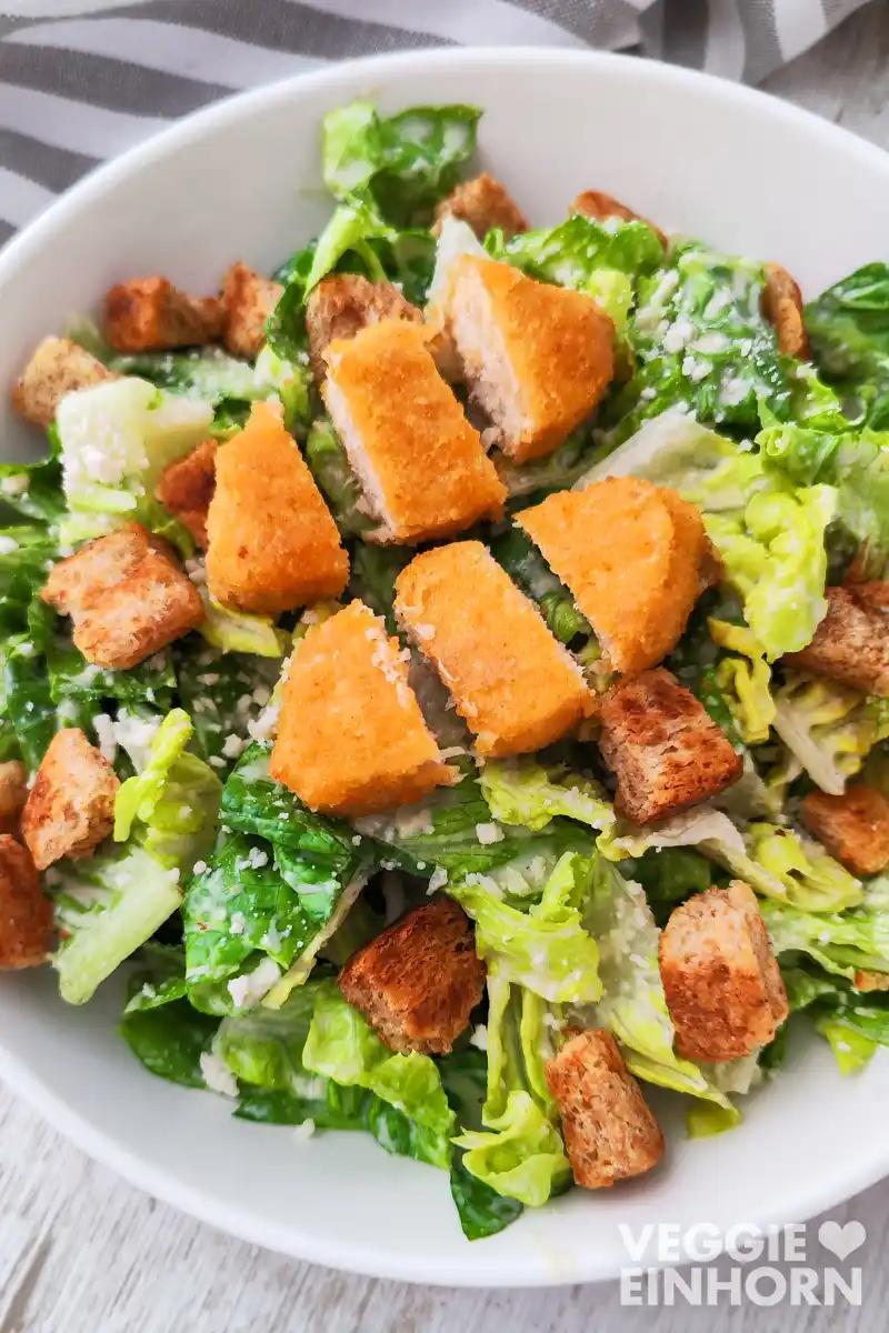 Ein veganer Caesar Salat aus Römersalat, Croutons und veganem Parmesan, mit klein geschnittenen veganen Nugget-Stücken.