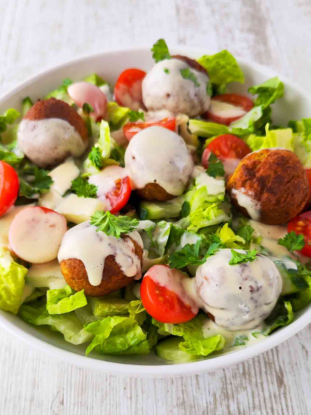 Falafel-Salat mit Romanasalat, Kirschtomaten, , Salatgurken und cremigem Dressing, serviert in einer weißen Schüssel.