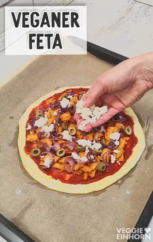 Pizza mit Tomatensauce, veganem Gyros, Zwiebeln und Oliven wird mit zerbröckeltem veganem Feta bestreut.
