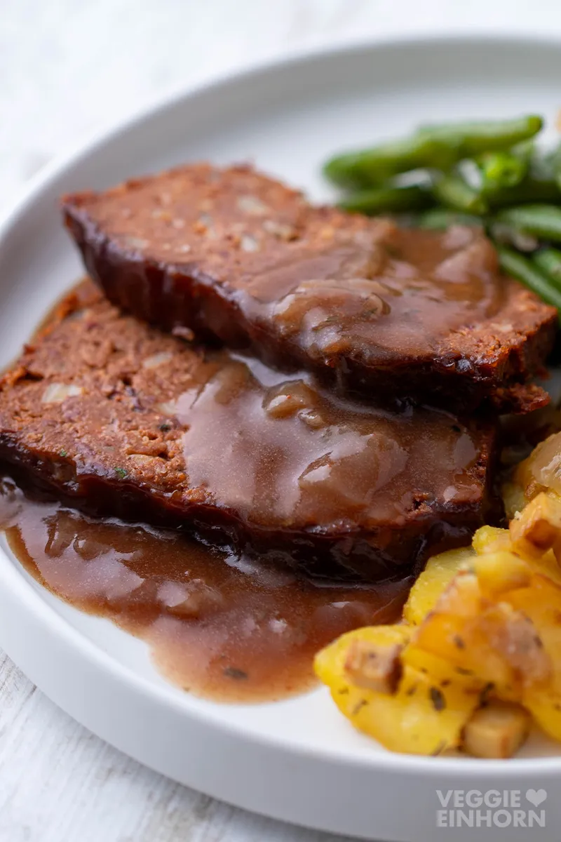Nahaufnahme von zwei Scheiben veganem Hackbraten aus Kidneybohnen und Linsen mit Zwiebelsauce auf einem Teller mit Bratkartoffeln und grünen Bohnen.