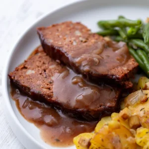 Zwei Scheiben veganer Hackbraten aus Kidneybohnen und Linsen mit Zwiebelsauce, grünen Bohnen und Bratkartoffeln auf einem weißen Teller serviert.