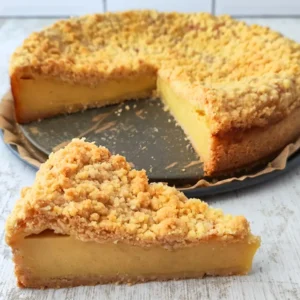 Ein veganer Käsekuchen mit Streuseln steht auf einer Backform, ein Stück wurde herausgenommen und liegt vorne auf dem Tisch.