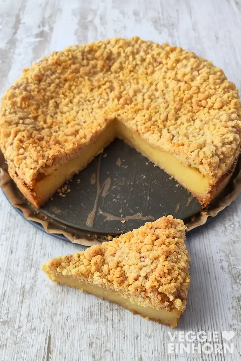 Ein veganer Käsekuchen mit Streuseln steht auf einem Springformboden, im Vordergrund liegt ein Stück des Kuchens.