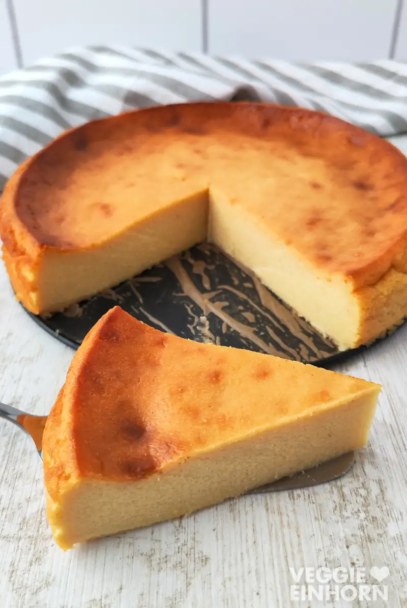 Ein Stück veganer Käsekuchen ohne Boden liegt auf einem Tortenheber und im Hintergrund steht der angeschnittene Kuchen auf einem Springformboden.