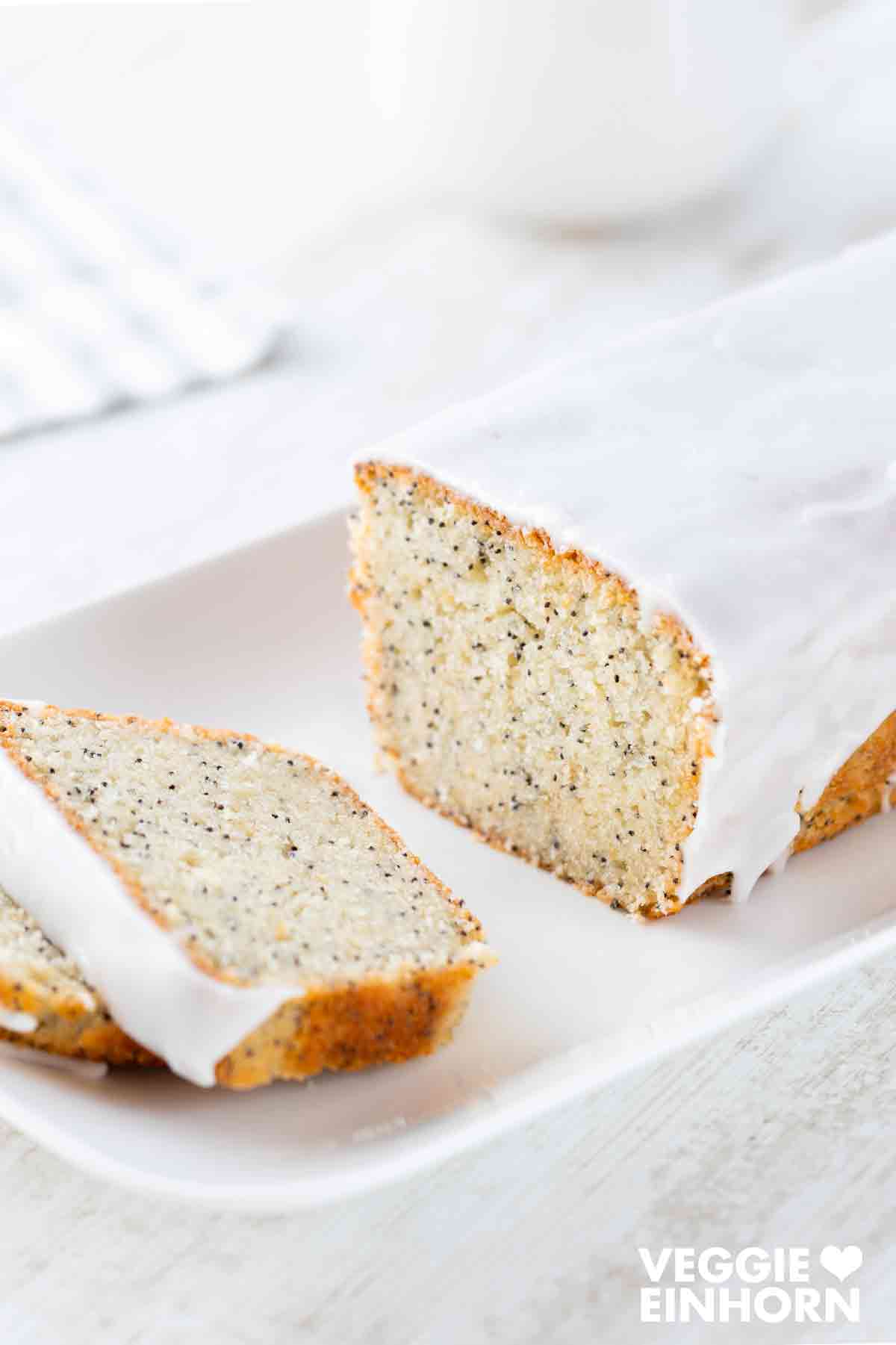Saftiger veganer Mohn-Zitronenkuchen mit Zuckerguss, angeschnitten auf einer weißen Kuchenplatte.