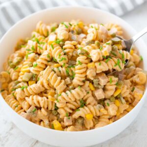 Eine weiße Schüssel mit cremigem veganem Nudelsalat aus Fusilli, veganer Fleischwurst, Erbsen, Mais, Mayo und Gewürzgurken, garniert mit frischem Schnittlauch. Ein Löffel hebt etwas Salat aus der Schüssel.