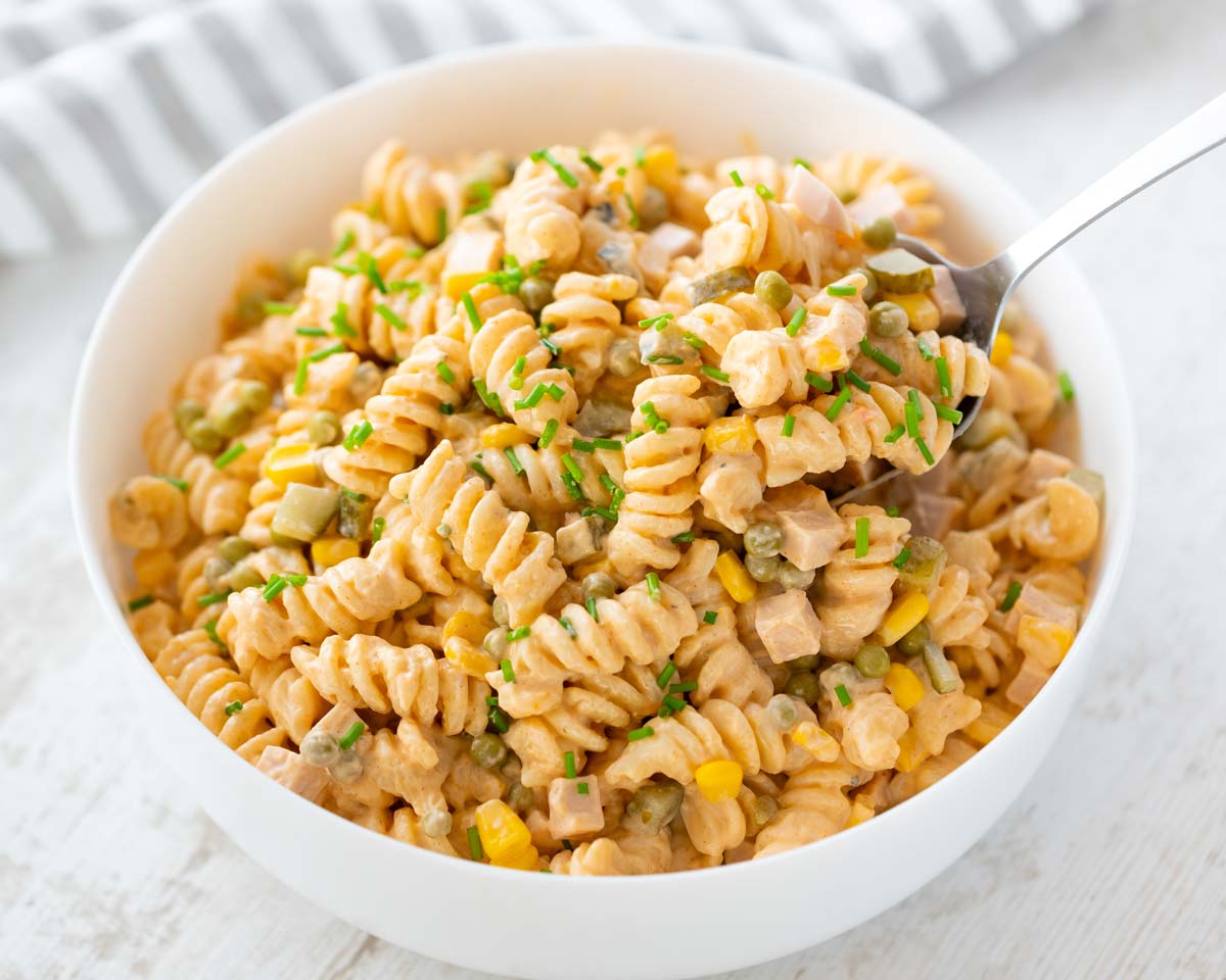 Eine weiße Schüssel mit cremigem veganem Nudelsalat aus Fusilli, veganer Fleischwurst, Erbsen, Mais, Mayo und Gewürzgurken, garniert mit frischem Schnittlauch. Ein Löffel hebt etwas Salat aus der Schüssel.